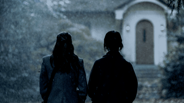 Two young women, seen from behind, face an unassuming house while standing in the rain