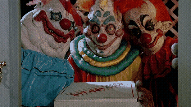 Three weird, lumpy clowns stand in a doorway holding a pizza box.