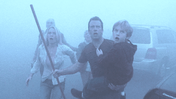 A man wielding a stick and carrrying a small child is backed up by several other people as they move through a foggy parking lot with worried expressions.