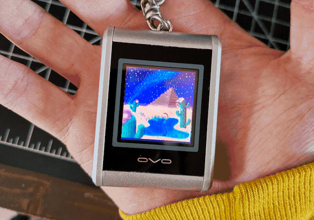 A tiny, silver device with a keychain attached to the top sitting in my hand. It has a small, square LCD screen on the front, which is displaying at a comically low resolution a piece of art I made.