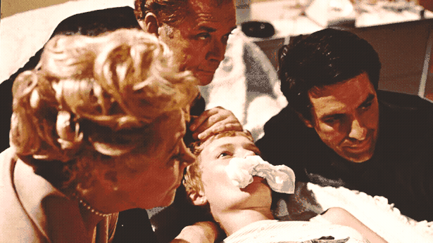 Mia Farrow looking unwell with a tissue stuck in her mouth is held down in bed by an old lady and two guys.