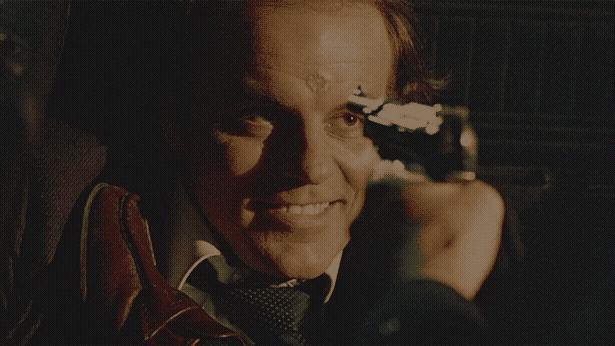 Michael Ironside, with a weird scar on his forehead, grins as a gun is pointed at his face.