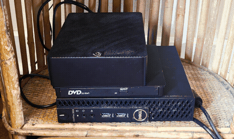 A small black computer box with a Dell logo, sitting in the bottom cubby of a bamboo shelf. On top of it are stacked an external DVD drive and a large hard drive.