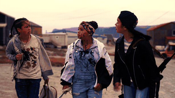 Three young girls walk across an Arctic village, all splattered with black goo. The right one is toting a rifle.