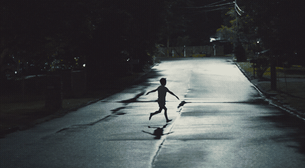 A kid runs down the street at night, backlit by wet asphalt.