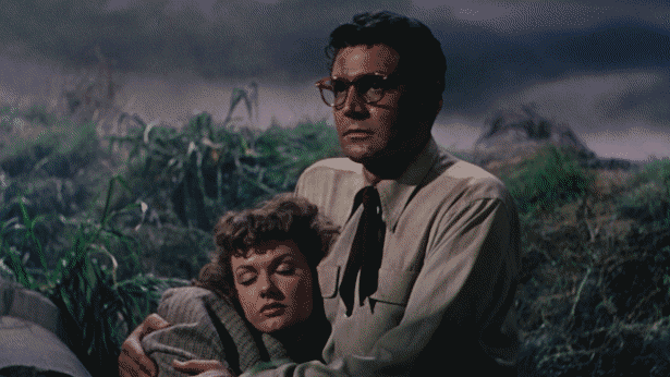 A weary-looking 1950s guy in glasses and a short tie holds a lady who is either passed-out or asleep in front of a foggy landscape of torn-up grass.
